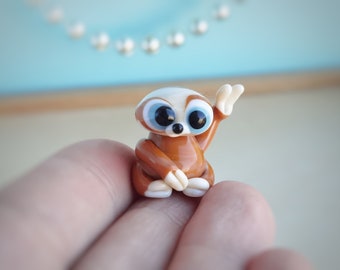 Puede incluir: Una pequeña figurita de vidrio de un perezoso marrón y blanco con grandes ojos azules. El perezoso está sentado con un brazo levantado.
