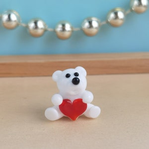 May include: A white glass bear figurine holding a red heart. The bear has black eyes and a small black nose.