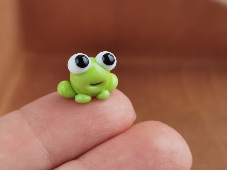 May include: A small green frog figurine with large black eyes and a white background. The frog is sitting on a finger.