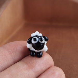 May include: A small black and white glass sheep figurine with large eyes. The sheep is standing on a person's finger.