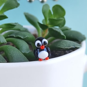 May include: A miniature glass penguin figurine featuring a glossy black body, a white belly, large white eyes with black pupils, and a small orange beak and feet. The penguin is positioned upright with a smooth, reflective surface finish.