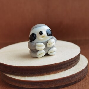 May include: A small glass figurine of a sloth, with a gray and white body, black eyes, and a white face. The figurine is sitting on a wooden platform.