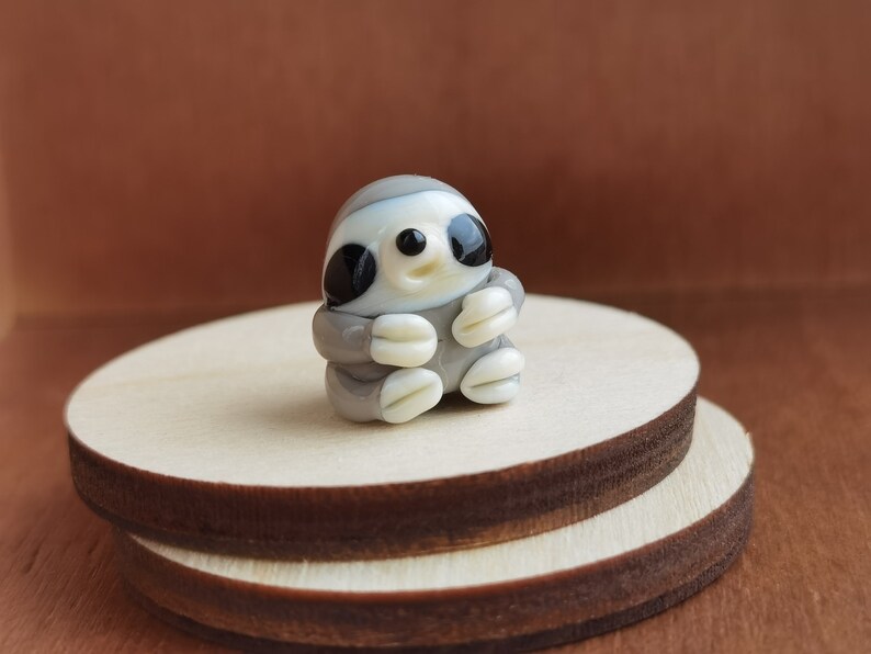 May include: A small glass figurine of a sloth, with a gray and white body, black eyes, and a white face. The figurine is sitting on a wooden platform.