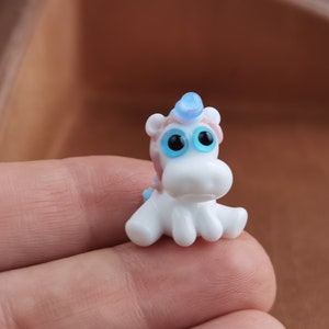 May include: A small white glass unicorn figurine with blue eyes and a blue horn. The unicorn is sitting on its hind legs with its front legs outstretched.