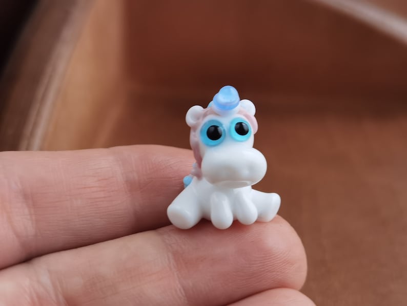 May include: A small white glass unicorn figurine with blue eyes and a blue horn. The unicorn is sitting on its hind legs with its front legs outstretched.