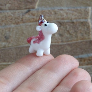 May include: A small white glass unicorn figurine with pink and silver accents. The unicorn has a horn, mane, and tail. The figurine is sitting on a hand.