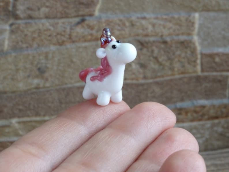Op de afbeelding: Een klein wit glazen eenhoornbeeldje met roze en zilveren accenten. De eenhoorn heeft een hoorn, manen en een staart. Het beeldje staat op een hand.