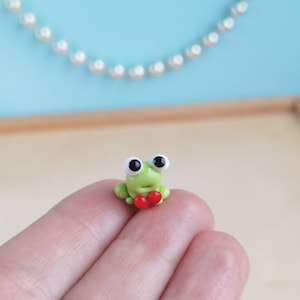 以下が含まれることがあります： 大きな黒い目をした小さな緑色のカエルの置物。手に赤いハートを持っている。