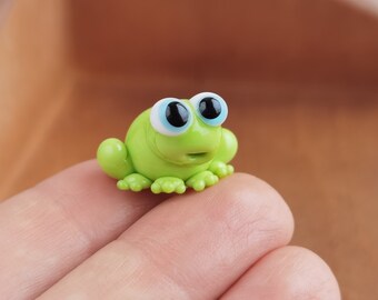 May include: A small green frog figurine with large blue eyes and a white rim around the eyes. The frog is sitting on a finger.