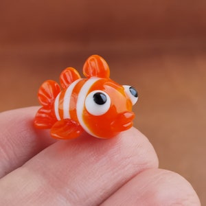 May include: A small, orange and white glass figurine of a clownfish. The fish has large, black eyes and a white stripe around its body.