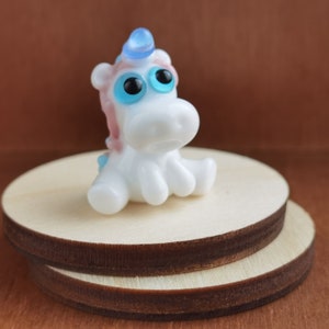 May include: A small white glass unicorn figurine with blue eyes and a blue horn. The unicorn is sitting on a wooden base.
