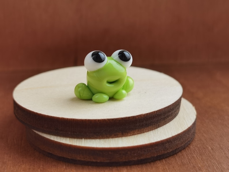 May include: A small green glass frog figurine with white eyes and black pupils. The frog is sitting on a wooden platform.