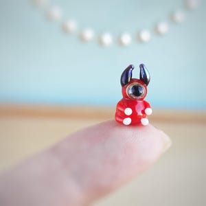Op de afbeelding: Een klein, rood glazen beeldje met een enkel zwart oog en twee kleine hoorns. Het beeldje zit op een vinger.
