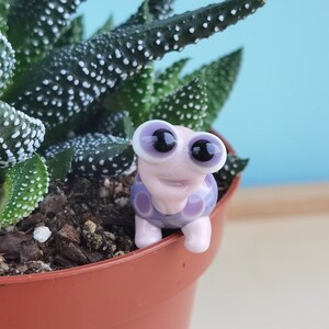 May include: A small purple and white ceramic turtle figurine with large black eyes and a smiling face. The turtle is sitting on the edge of a terracotta pot filled with soil and a green succulent plant.