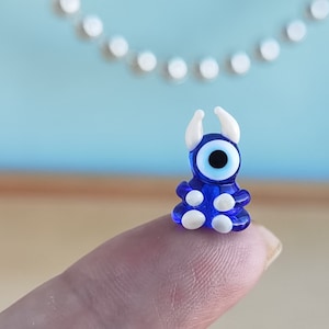 May include: A small blue glass figurine with a white evil eye and white dots. The figurine is sitting on a finger.