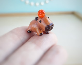 May include: A small, hand-painted glass figurine of a capybara wearing a bright orange hat. The figurine is brown and has black eyes and nose.