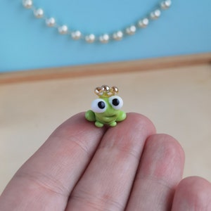 以下が含まれることがあります： 金色の冠をかぶった小さな緑色のカエルの置物。カエルは大きな白い目と小さな笑顔をしています。置物は手に乗っています。