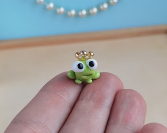 May include: A small green frog figurine wearing a gold crown. The frog has large white eyes and a small smile. The figurine is sitting on a hand.