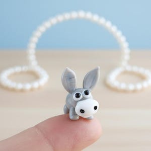 May include: A miniature gray donkey figurine with large ears, black eyes, and a white muzzle. The donkey is perched on a fingertip, with a blurred pearl necklace in the background. The figurine is a small, detailed animal sculpture.
