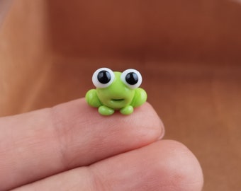 May include: A small green frog figurine with large white eyes and black pupils. The frog is sitting on a finger.