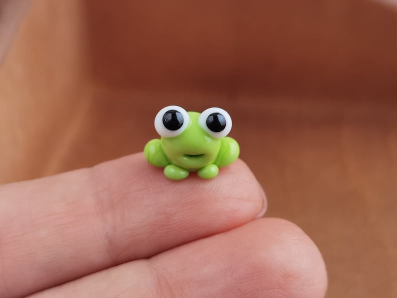May include: A small green frog figurine with large white eyes and black pupils. The frog is sitting on a finger.