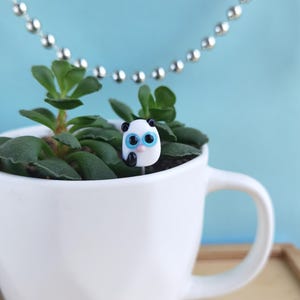 Puede incluir: Una pequeña figura de panda de cristal con grandes ojos azules y orejas negras está colocada dentro de una planta suculenta verde en una taza de cerámica blanca. Una cadena de bolas plateada cuelga al fondo sobre un fondo azul claro.
