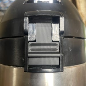 May include: Close-up of a stainless steel and black insulated thermos. The black lid features a hinged locking mechanism. The thermos has a brushed metal finish and a black plastic top.