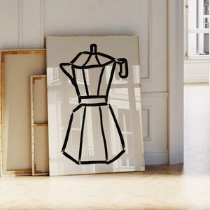 May include: A framed art print featuring a minimalist black line drawing of a stovetop espresso maker. The artwork is set against a neutral background with a gold frame. Other framed canvases lean against the wall. The overall theme is home decor.