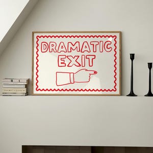 Puede incluir: Un cuadro enmarcado con las palabras "DRAMATIC EXIT" en rojo y una mano que señala. El cuadro tiene un fondo blanco y un borde rojo festoneado, en un marco marrón claro. Dos candelabros negros a la derecha.