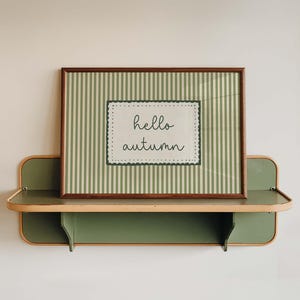 May include: A rectangular framed print featuring a sage green and white vertical striped background with a central white rectangular label containing the text hello autumn in black script, resting on a sage green wall-mounted shelf.