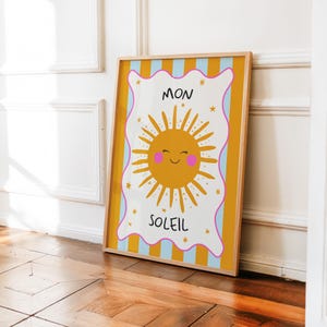 Puede incluir: Una impresión artística enmarcada con una ilustración de sol sonriente y las palabras "MON SOLEIL". El sol es amarillo con mejillas rosas y líneas radiantes. El fondo tiene rayas verticales amarillas, azules y blancas.