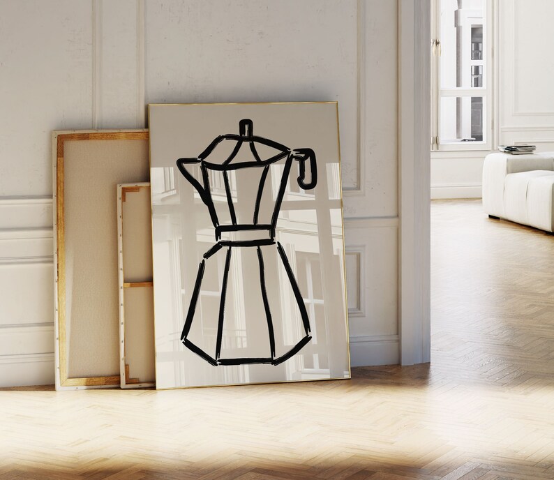 May include: A framed art print featuring a minimalist black line drawing of a stovetop espresso maker. The artwork is set against a neutral background with a gold frame. Other framed canvases lean against the wall. The overall theme is home decor.