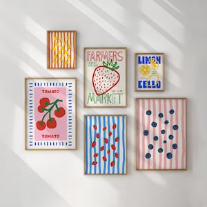 May include: A collection of framed art prints featuring colorful fruit illustrations. Prints include lemons, strawberries, tomatoes, and blueberries. One print reads "Farmers Market" with a strawberry graphic. The prints are displayed on a white wall.