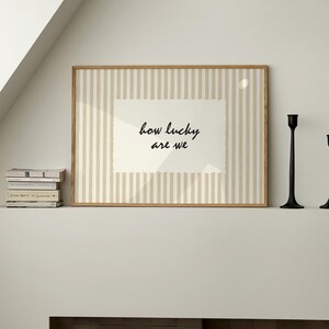 Op de afbeelding: Een ingelijste print met de tekst "how lucky are we" in zwart schrift. De achtergrond heeft verticale beige en witte strepen. De lijst is gemaakt van lichtbruin hout. Twee zwarte kandelaars staan rechts.