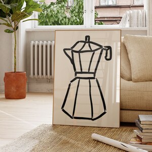 May include: A framed art print featuring a black line drawing of a stovetop espresso maker on a cream-colored background. The artwork is displayed in a living room setting, leaning against a wall near a window and a beige sofa.