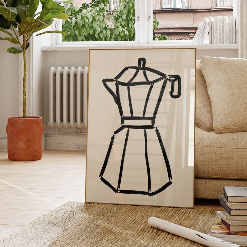 May include: A framed art print featuring a black line drawing of a stovetop espresso maker on a cream-colored background. The artwork is displayed in a living room setting, leaning against a wall near a window and a beige sofa.