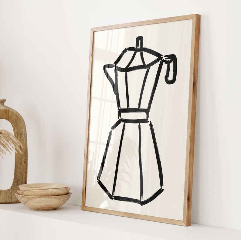 May include: A framed art print featuring a black and white illustration of a stovetop espresso maker. The artwork is a minimalist design with bold lines on a cream-colored background, set within a natural wood frame. The print is leaning against a white wall.