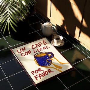 以下が含まれることがあります： 白い背景に赤い文字で「Un cafe con leche por favor」と書かれた額装されたプリント。プリントには、青いコーヒーカップを黄色い手で持った漫画のイラストが描かれている。