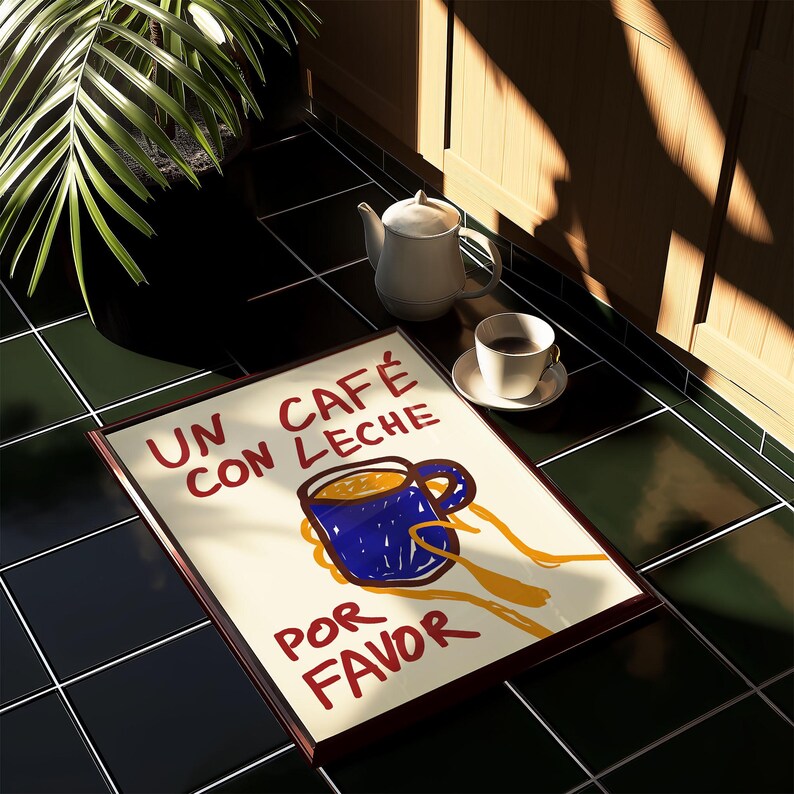 以下が含まれることがあります： 白い背景に赤い文字で「Un cafe con leche por favor」と書かれた額装されたプリント。プリントには、青いコーヒーカップを黄色い手で持った漫画のイラストが描かれている。