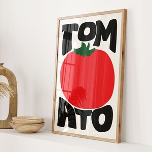 May include: A framed print with the word "TOMATO" in bold black letters. The word "TO" is stacked on top of the word "MATO". A red tomato with a green stem is centred between the words.