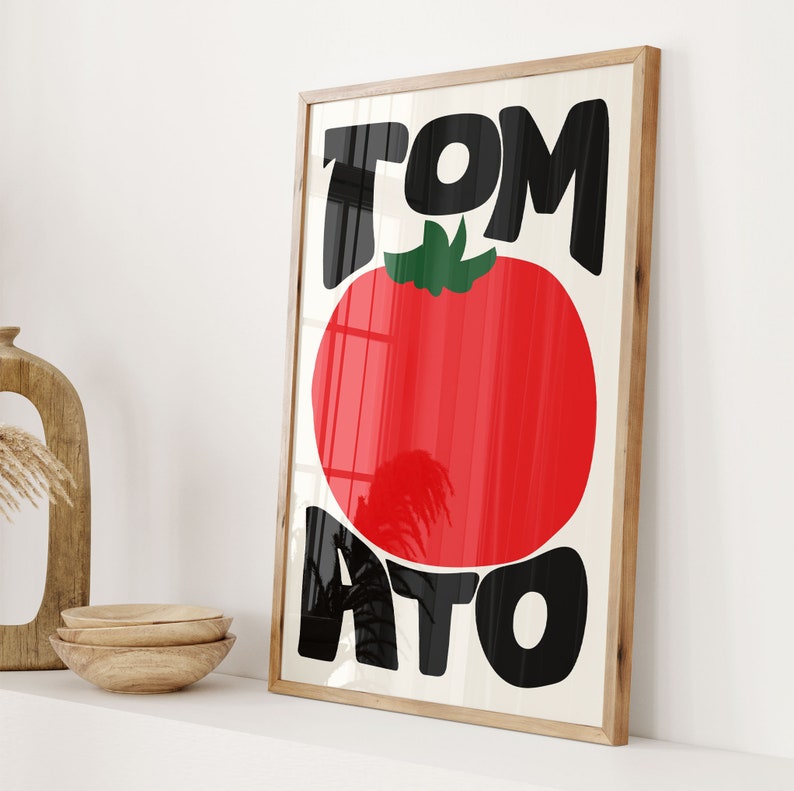 May include: A framed print with the word "TOMATO" in bold black letters. The word "TO" is stacked on top of the word "MATO". A red tomato with a green stem is centred between the words.