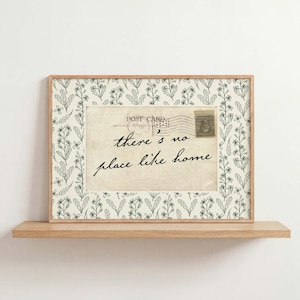 May include: A framed postcard with the text "there's no place like home." The postcard is cream-coloured with a vintage stamp and postmark. The frame is light wood with a floral pattern border, displayed on a wooden shelf.