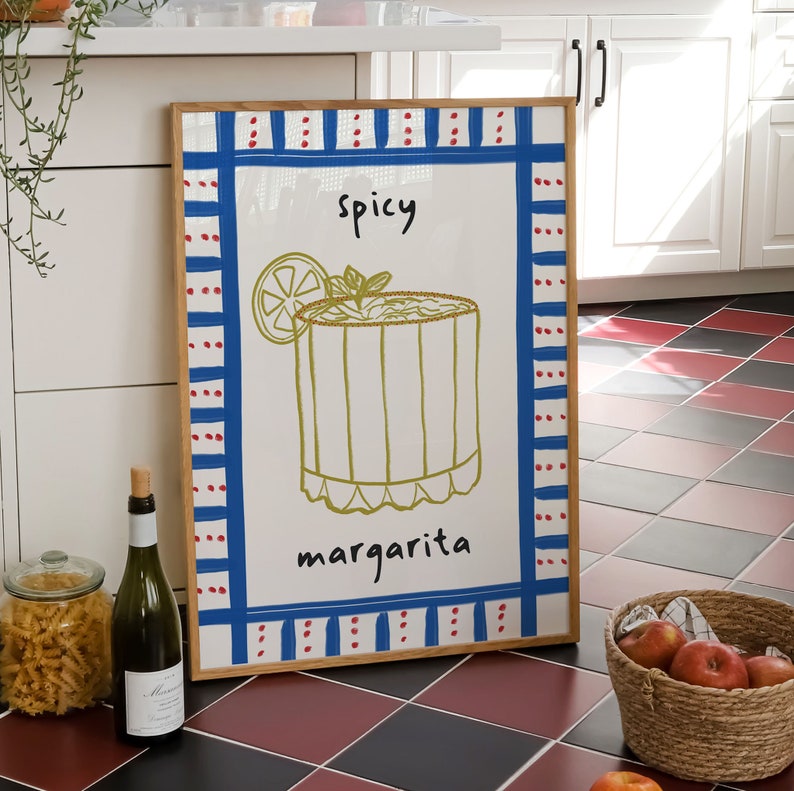 May include: A framed art print featuring a margarita cocktail illustration. The artwork has a white background with a blue and red border. The words "spicy margarita" are written on the print. A bottle of wine and a basket of apples are in the foreground.