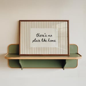 May include: A framed print with the text "there's no place like home" sits on a green and tan metal shelf. The print has a brown frame and a striped background. The shelf is mounted on a white wall.