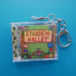 May include: A clear plastic keychain with a Stardew Valley design. The keychain has a rectangular shape with a printed image of the game's logo and characters. The keychain includes a silver clasp and a key ring.