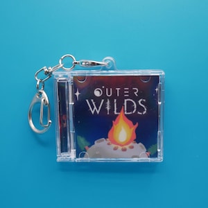 Può includere: Un portachiavi in plastica trasparente con una custodia per CD in miniatura con le parole "OUTER WILDS" e una grafica di falò. Il portachiavi ha un fermaglio e un anello argentati, su sfondo blu.