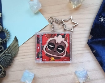 May include: A square, clear plastic keychain case featuring an illustration of a cartoon lamb with large eyes and a small bell on a red background. The keychain includes a silver-toned metal clasp and a star-shaped charm attached to the top.