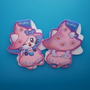 Puede incluir: Dos pegatinas troqueladas con un personaje felino con sombrero de seta y peto. Las pegatinas son principalmente rosas y azules, decoradas con motivos florales. El texto "@yai_dibuja" está impreso en la parte superior de cada pegatina.