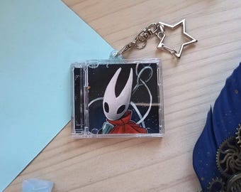 May include: A square clear plastic keychain featuring a miniature CD case design. Inside the case is an illustration of a white horned character with black eyes wearing a red cloak against a dark background. It has a silver star-shaped clasp.