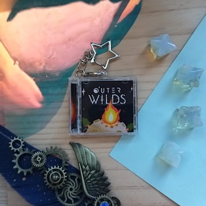 Puede incluir: Un llavero cuadrado de plástico transparente con una réplica en miniatura de una caja de CD de Outer Wilds. La portada muestra el título del juego en texto blanco sobre un gráfico de una fogata en un planeta. Incluye un cierre metálico en forma de estrella.